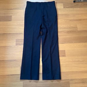 May include: Dark navy blue trousers with a high waist and wide legs. The pants are made of a smooth fabric and have a classic, tailored design. The trousers are displayed on a light brown wooden surface.