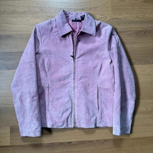 May include: A light pink suede jacket with a collared neckline and a full-length zipper. The jacket has two front pockets and long sleeves. The jacket is displayed on a wooden surface.
