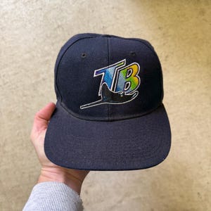 May include: Navy blue baseball cap with a flat brim. The cap features an embroidered logo with the letters "TB" in a gradient of blue, green, and yellow, with a stylized ray design. The cap is a fitted style.