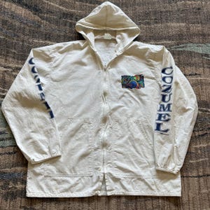 May include: Off-white hooded zip-up jacket with the word "CoZUMEL" printed in navy blue on the sleeves. A colorful graphic patch is on the chest. The jacket has a full zipper and two front pockets.