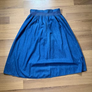 May include: A medium-wash blue denim skirt featuring a high-waisted design with a wide, gathered elastic waistband. The skirt has a full, A-line silhouette that flares out from the waist down to the hem, creating soft vertical pleats throughout.