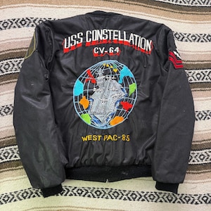 May include: A black bomber jacket featuring back embroidery that reads USS CONSTELLATION CV-64 and WEST PAC-85. The design includes a globe graphic with a central aircraft carrier, airplanes, and colorful landmasses in red, blue, orange, and green.
