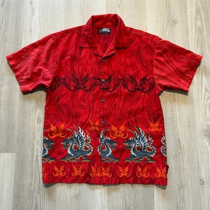 Puede incluir: Camisa roja de manga corta con un patrón arremolinado y cierre de botones. Presenta diseños tribales negros y gráficos de dragones con acentos grises y blancos. La etiqueta dice "Pineapple Connection".