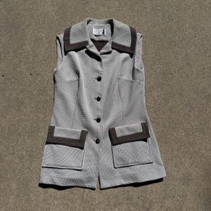 May include: A vintage sleeveless vest in a black and white checkered pattern. The vest features a notched collar and two front pockets, both trimmed with a dark brown fabric. It has a button-down closure with black buttons.