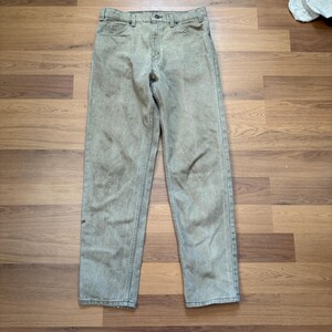 May include: A pair of olive green denim jeans. The jeans have a classic five-pocket design with a button and zipper closure. The pants are laid flat on a wooden surface.