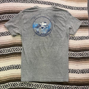 May include: A heather gray short-sleeve t-shirt featuring a circular graphic on the back. The design depicts a mountain peak, blue sky, and pine trees framed by two crossed ice axes, with the text GRAND TETONS printed in dark blue lettering.