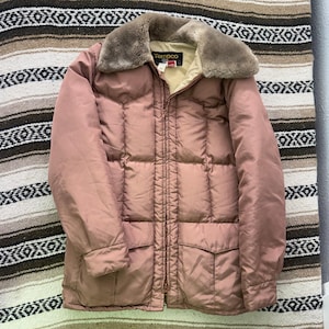 May include: A dusty rose quilted puffer jacket featuring a brown faux fur collar, a full-length front zipper, and two large front pockets with flaps. The interior lining is beige, and a black Tempco brand label is visible at the neckline.
