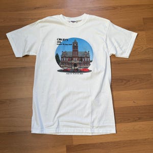 Puede incluir: Camiseta blanca con un gráfico de un edificio de ladrillo con una torre de reloj dentro de un círculo azul. El texto "COLBY'S 10th Cruise To The Past" y la fecha "June 13, 14, & 15, 2003" están impresos en la camiseta.