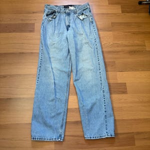 May include: Light blue denim jeans with a relaxed fit. The jeans feature a classic five-pocket design, a button and zip closure, and belt loops. The denim has a faded appearance with some wear and tear, giving them a vintage look.