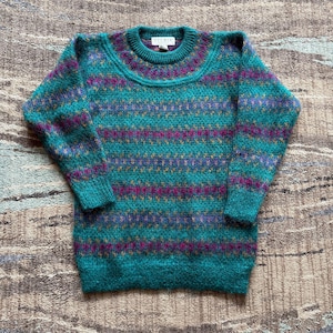 May include: A long-sleeved crew neck sweater featuring a textured knit pattern in shades of teal, magenta, and purple. The garment displays horizontal bands of intricate geometric designs across the chest, sleeves, and body, with a ribbed collar.