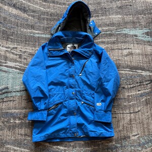 以下が含まれることがあります： 鮮やかな青色のフード付きジャケット、内側はグレー。 ジャケットはジッパー開閉、複数のポケット、調節可能な袖口付き。 胸元にThe North Faceのロゴが見えます。 アウトドア活動に適しています。