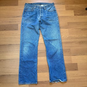 May include: Faded blue denim jeans with a button fly and classic five-pocket design. The jeans show signs of wear and fading, with a straight leg cut. The denim appears to be a medium weight, suitable for everyday wear.