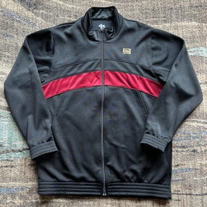 May include: Black track jacket with a full-length zipper, a stand-up collar, and a burgundy stripe across the chest. The jacket has a gold logo on the chest and ribbed cuffs and hem. A sporty, casual jacket.
