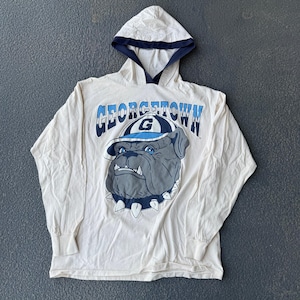 May include: Cream-coloured long-sleeved hoodie with a Georgetown Bulldog graphic. The graphic includes the word "Georgetown" in blue and white, and a bulldog wearing a hat. The hood has a navy blue trim.