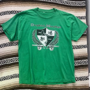 May include: A bright green short-sleeve t-shirt featuring a graphic print of a shield with a hammer and lion emblem. The text Dropkick Murphys is printed above the shield, which is flanked by laurel branches and two cannons at the base.