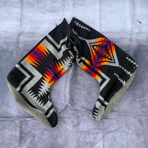 Pode incluir: Par de botas de lã pretas e cinzentas com um padrão geométrico em laranja, roxo e branco. As botas têm uma sola de camurça cinzenta e um fecho de cordão. O design é inspirado na arte nativa americana.