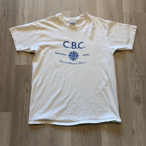 Puede incluir: Camiseta blanca con un logotipo azul y blanco para la Canadian Broadcasting Corporation (CBC). El logotipo presenta un globo terráqueo estilizado con el texto "C.B.C." encima y "Televisión" y "Radio" a cada lado. El texto "¡Isla del Príncipe Eduardo!" está debajo del logotipo.