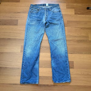 May include: Faded blue denim jeans with a button fly. The jeans have a classic five-pocket design and a straight leg cut. The denim shows signs of wear and fading, giving them a vintage look. The jeans are laid flat on a wooden surface.