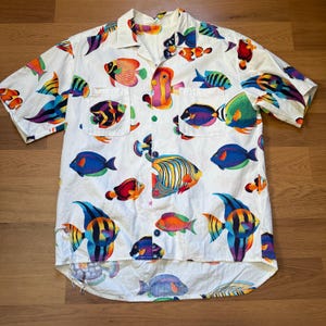 Può includere: Camicia bianca con bottoni con un vivace motivo a pesce. La camicia a maniche corte presenta illustrazioni di pesci colorati nelle tonalità di blu, arancione, giallo e verde. Due tasche anteriori e bottoni verdi completano il design. Un capo a tema tropicale.
