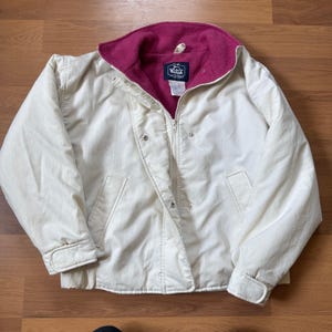 May include: A cream-colored jacket featuring a vibrant magenta fleece lining and a high collar. The jacket includes a front zipper closure, snap buttons, and two side pockets. A label inside the collar reads The Woolrich Woman.