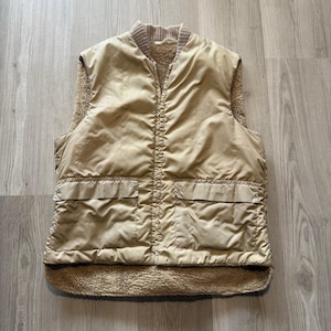 May include: A tan-colored, quilted vest with a fleece lining and two front pockets. The vest has a ribbed collar and a full-length zipper. The vest is a neutral color and appears to be made of a durable material.