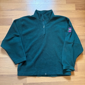 May include: A dark teal fleece pullover with a quarter-zip and a high collar. The long-sleeved top features a small patch on the upper left sleeve with text and a logo. The hemline has a drawstring. The garment is made of a soft, warm material.