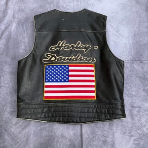 May include: Black leather vest with the Harley-Davidson logo and an American flag patch. The vest has a classic biker style with a quilted lower section and a simple collar.