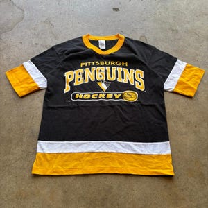 Könnte beinhalten: Schwarzes Pittsburgh Penguins Hockey-Trikot mit gelben und weißen Akzenten. Das Trikot zeigt den Teamnamen und das Logo in Gelb und Weiß, mit einem gelben Kragen und Ärmelbündchen. Das Wort "HOCKEY" steht unter dem Teamnamen.