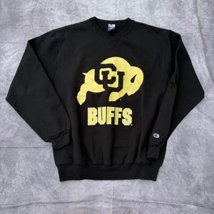 Może przedstawiać: Czarna bluza z okrągłym dekoltem z żółtym logo CU Buffaloes i napisem "BUFFS" w żółtych literach. Bluza ma długie rękawy i logo Champion na lewym rękawie.