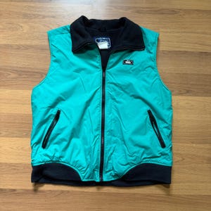 May include: A bright teal sleeveless vest featuring a black fleece collar and a matching black fleece hem. The garment includes a full-length black front zipper, two slanted zippered side pockets, and a small black rectangular logo patch.
