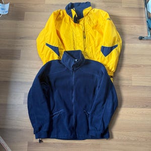 May include: Two jackets: a bright yellow waterproof jacket with navy blue detailing and a navy blue fleece jacket. The yellow jacket has a hood and pockets. The fleece jacket has a full zip and two side pockets. Both jackets are laid flat.
