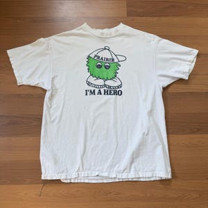 May include: White t-shirt featuring a green cartoon character wearing a cap with the word "Prairie" and the text "I'M A HERO". The graphic is in black and green. The shirt is made of a soft, comfortable material.
