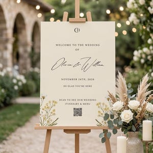 May include: A cream wedding sign on a wooden easel, with the text "Welcome to the Wedding of Olivia & William, November 24th, 2026." Floral decorations and a QR code are included. A vase of white roses and candles are in the foreground.
