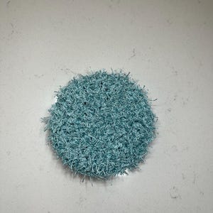 May include: A round, light blue crocheted scrubby. The scrubby is made of a textured, looped yarn, and is approximately 4 inches in diameter. The color is a soft, muted blue, and the overall appearance is fluffy and handmade.