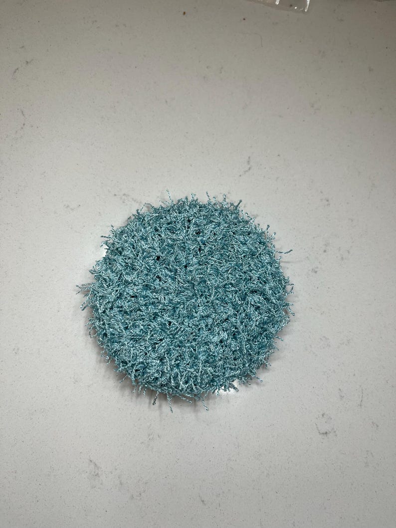 May include: A round, light blue crocheted scrubby. The scrubby is made of a textured, looped yarn, and is approximately 4 inches in diameter. The color is a soft, muted blue, and the overall appearance is fluffy and handmade.