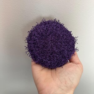 May include: A round, deep purple scrubby held in a hand. The scrubby has a textured, looped appearance, with small strands extending from the surface. The background is a plain, light gray wall.