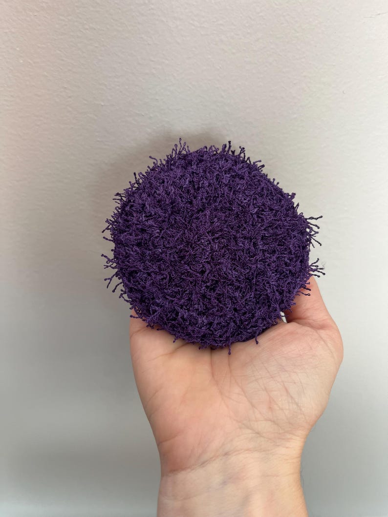 May include: A round, deep purple scrubby held in a hand. The scrubby has a textured, looped appearance, with small strands extending from the surface. The background is a plain, light gray wall.