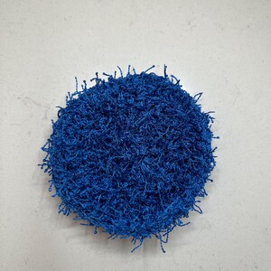 May include: A round, textured, blue scrubby. The scrubby is made of a looped, fibrous material. The edges of the scrubby have loose, frayed strands. The background is a plain, light gray surface.