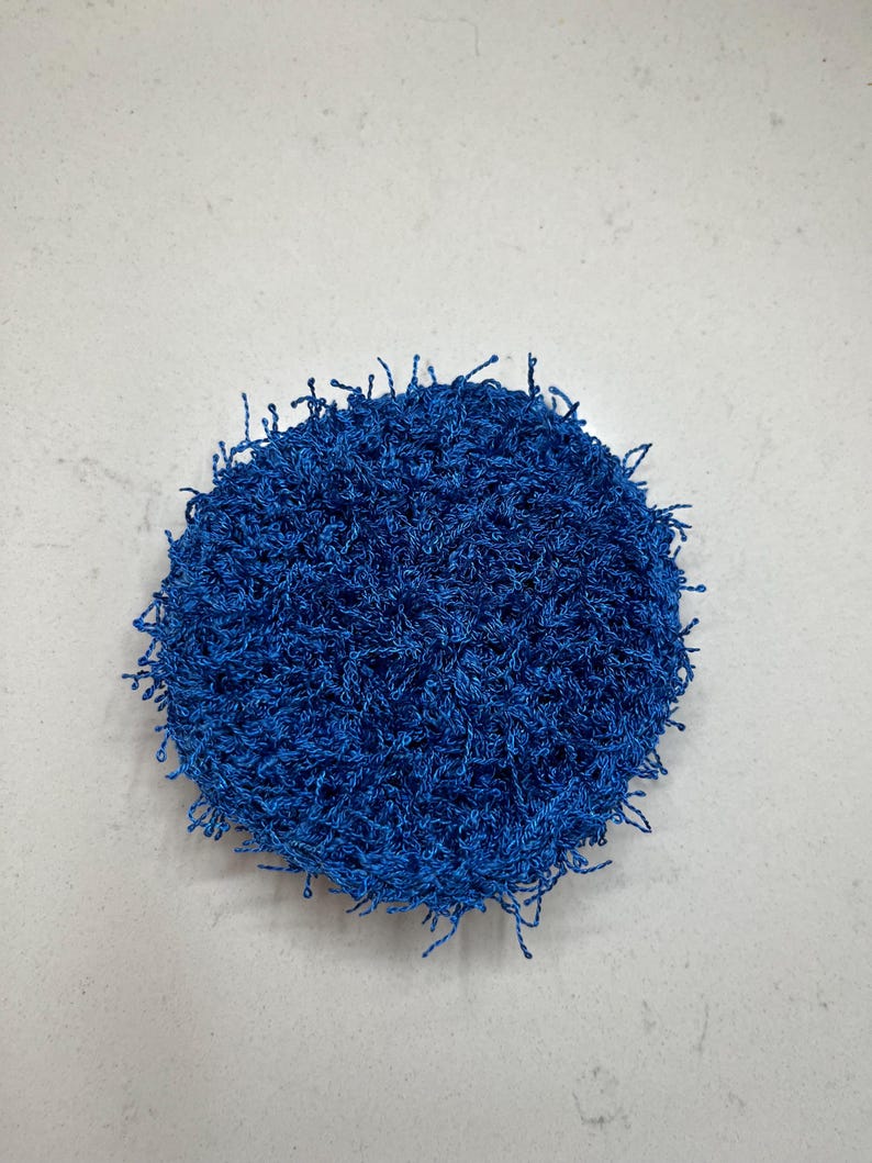 May include: A round, textured, blue scrubby. The scrubby is made of a looped, fibrous material. The edges of the scrubby have loose, frayed strands. The background is a plain, light gray surface.
