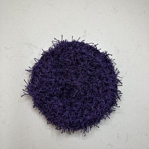 May include: A round, deep purple crocheted item with a textured surface and fringe-like edges. The item is centered on a light gray surface. The crocheted item appears to be handmade.