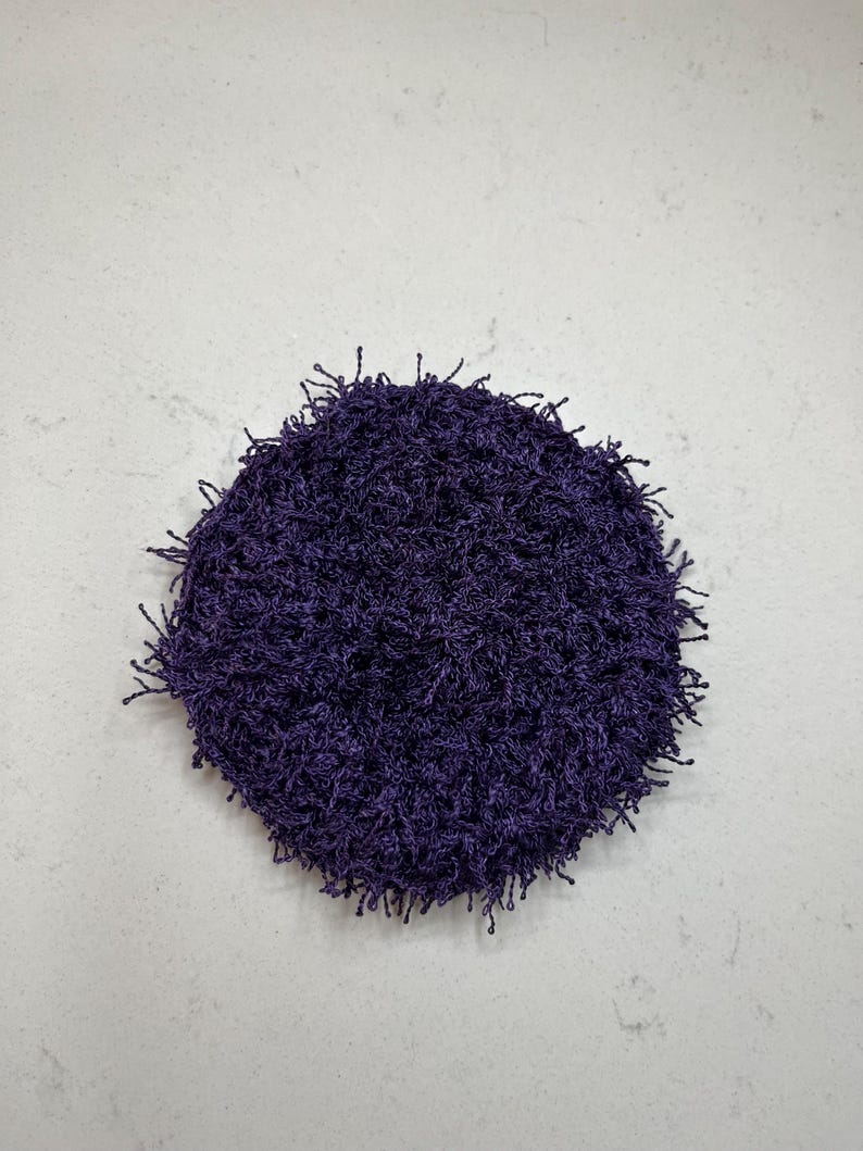 May include: A round, deep purple crocheted item with a textured surface and fringe-like edges. The item is centered on a light gray surface. The crocheted item appears to be handmade.
