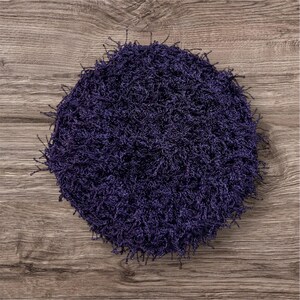 May include: A round, dark purple, shaggy rug with a textured surface. The rug is made of a soft, looped material and has a fringe around the edges. The rug is placed on a wooden surface.