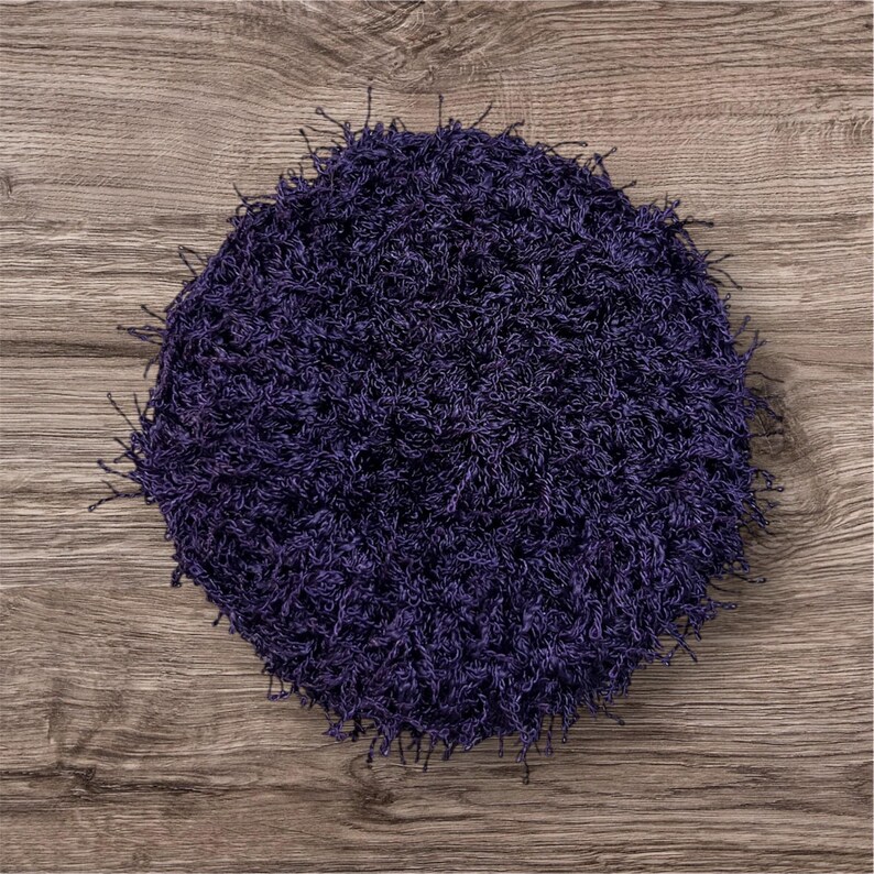May include: A round, dark purple, shaggy rug with a textured surface. The rug is made of a soft, looped material and has a fringe around the edges. The rug is placed on a wooden surface.