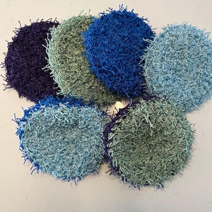 May include: A collection of seven handmade, circular scrubbies in various shades of blue, green, and purple. The scrubbies have a textured, looped appearance, with fringe-like edges. They are arranged in a circular pattern on a white surface.