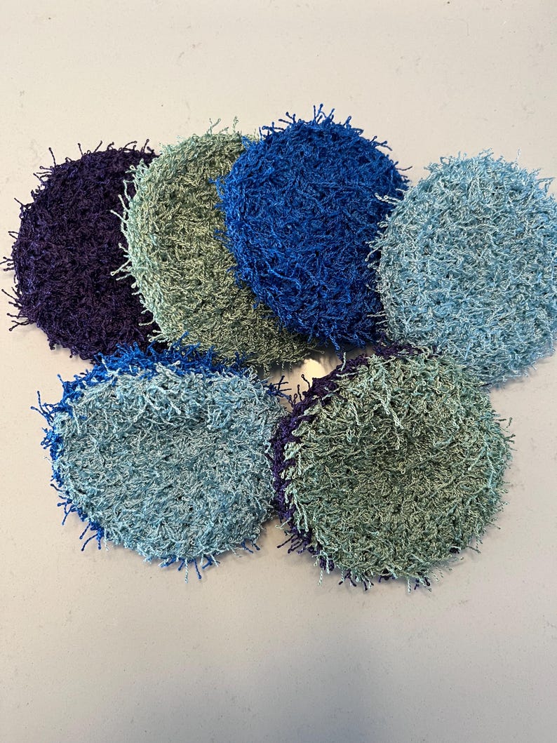 May include: A collection of seven handmade, circular scrubbies in various shades of blue, green, and purple. The scrubbies have a textured, looped appearance, with fringe-like edges. They are arranged in a circular pattern on a white surface.