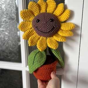 May include: A crocheted sunflower with a smiling face, yellow petals, brown center, green leaves, and an orange pot.