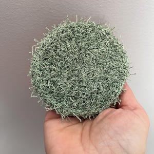 May include: A round, light green crocheted scrubby held in a hand. The scrubby is made of a textured yarn, with a dense, looped appearance. The color is a muted, soft green, with a slightly uneven texture.
