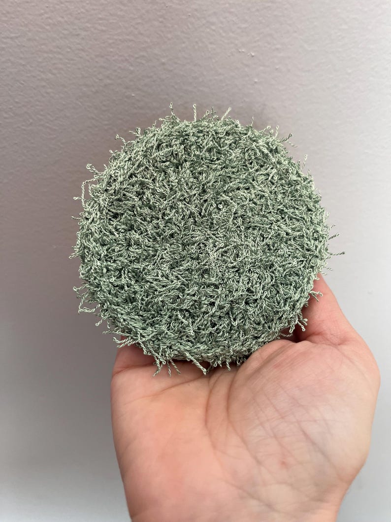 May include: A round, light green crocheted scrubby held in a hand. The scrubby is made of a textured yarn, with a dense, looped appearance. The color is a muted, soft green, with a slightly uneven texture.