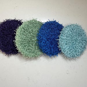 May include: Four round, textured scrubbies in shades of purple, green, blue, and teal. The scrubbies have a fuzzy, looped texture and are arranged in a row against a light background. These are likely for kitchen or bathroom use.