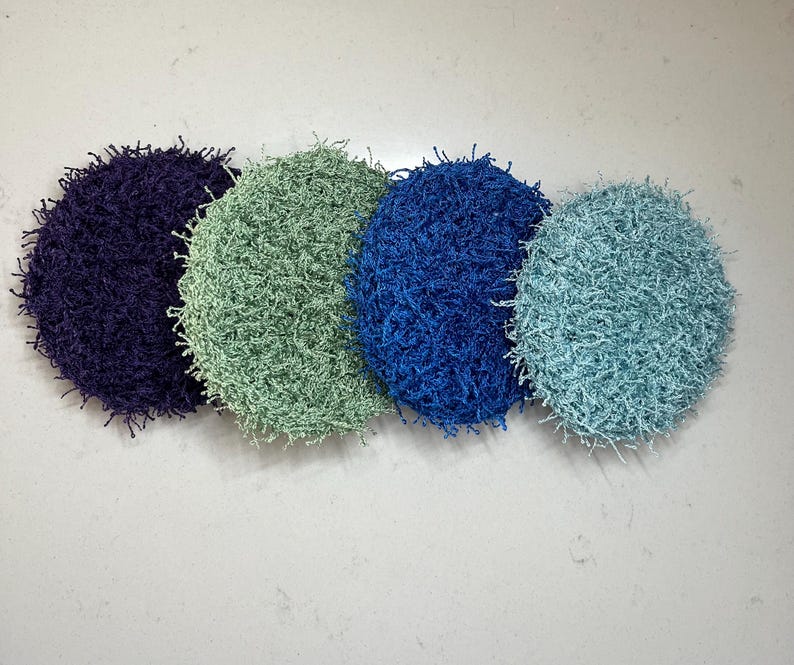 May include: Four round, textured scrubbies in shades of purple, green, blue, and teal. The scrubbies have a fuzzy, looped texture and are arranged in a row against a light background. These are likely for kitchen or bathroom use.
